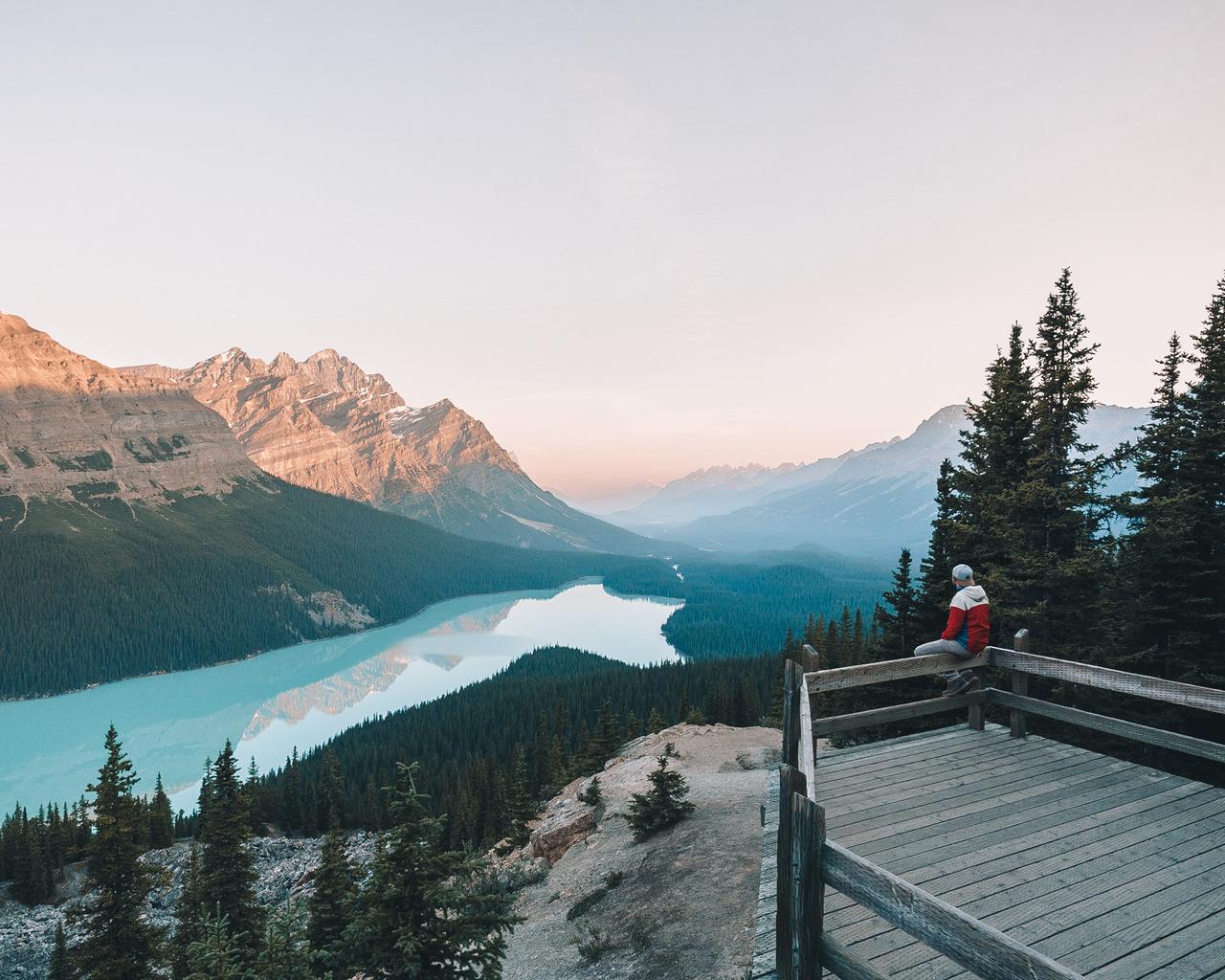 Person sitting on a balcony ledge looking out to a lake surrounded by mountains.